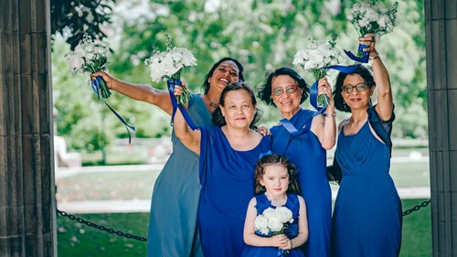 Bridesmaids in blue raising white bouquets under a stone arch