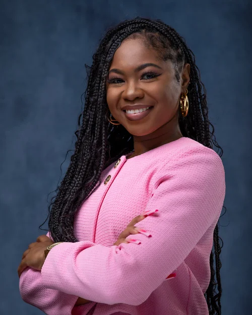 Woman with braids in a pink jacket, arms crossed, on a blue backdrop