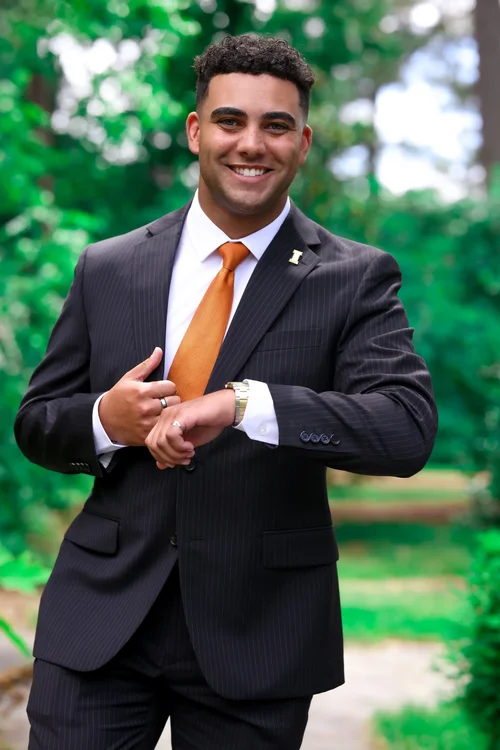Smiling man in a pinstripe suit and orange tie adjusting his cuff in a garden
