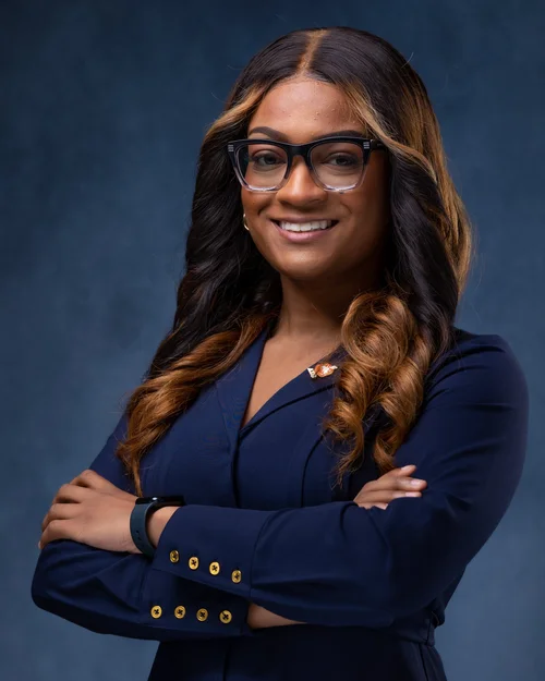 Woman in glasses and a navy blazer, arms crossed, on a blue backdrop