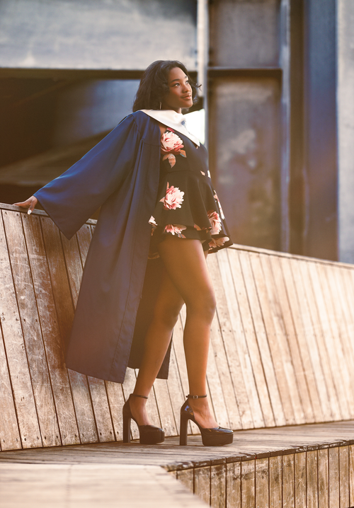 Graduate in an open navy gown leaning on a wooden bridge rail