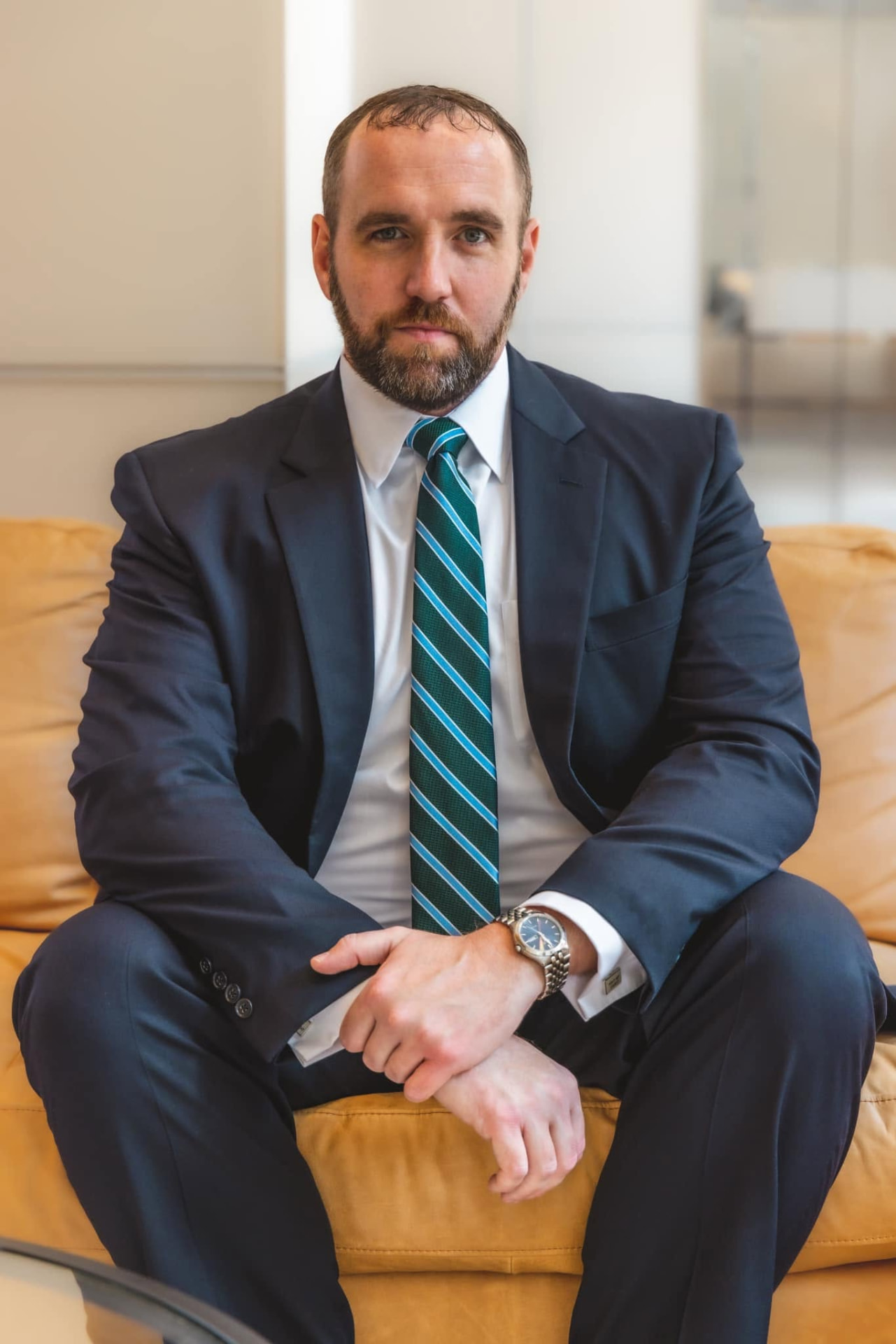 Man in a navy suit and striped tie seated on a tan sofa