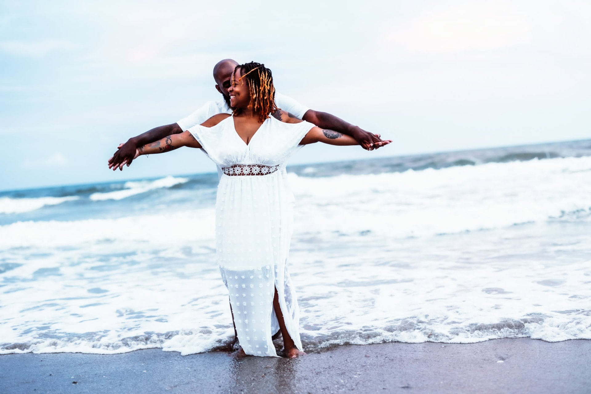 Couple in white embracing at the water's edge, arms wide open — Intimate wedding photography by Claudia Quiana