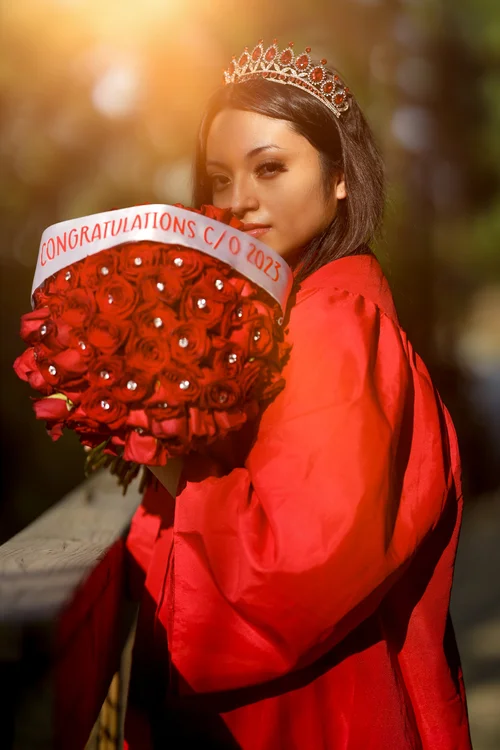 Graduate in a red gown and tiara holding a bouquet of red roses in golden light