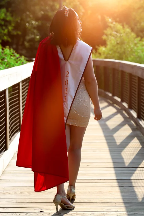 Graduate in a red gown and sash walking a sunlit bridge