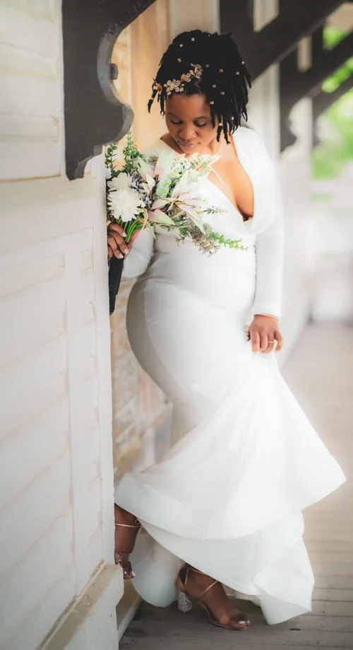 Bride in a fitted white gown leaning on a porch post with her bouquet