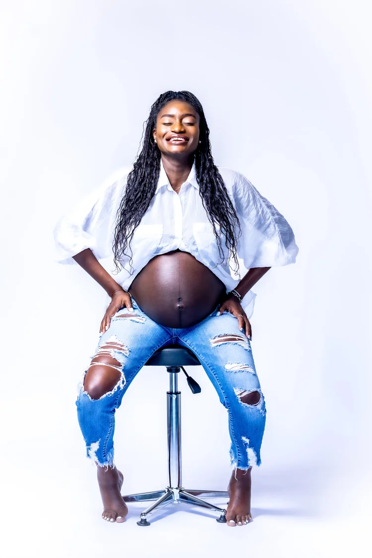 Smiling mother-to-be in a white shirt and ripped denim on a studio stool
