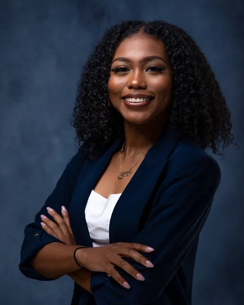 Smiling woman in a navy blazer and gold necklace on a blue backdrop